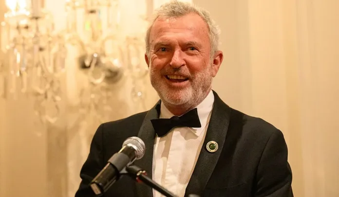 Sam Neill smiles while speaking at a microphone during a formal event, wearing a black tuxedo and bow tie, as Sam Neill tributes honour the acclaimed actor's legacy.