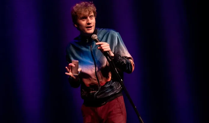 James Acaster performing stand-up comedy on stage with a microphone