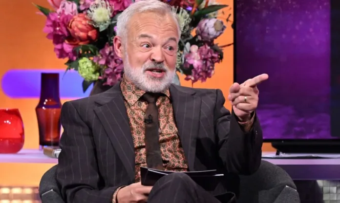 Graham Norton | The Graham Norton Show Series 34 Graham Norton wearing a dark pinstripe suit, seated on the set of The Graham Norton Show and pointing while speaking.