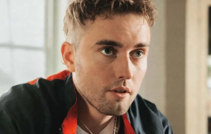 Close-up portrait of Sam Fender wearing a black jacket with red detailing, ahead of his 2027 stadium tour.